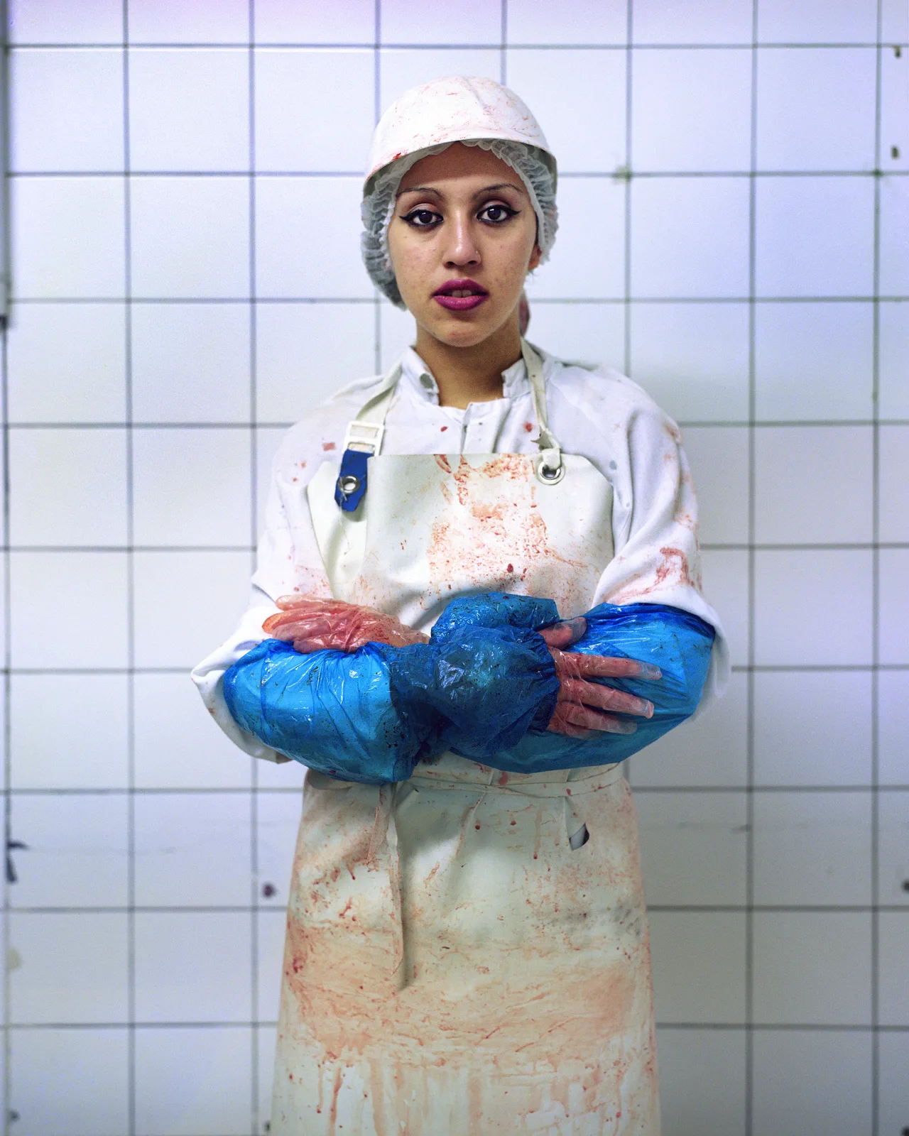 Portrait d'une jeune ouvrière d'abattoir, bras croisés devant un mur de carrelage blanc. Charlotte, tablier taché de sang, manchettes bleues, rouge à lèvres soutenu.