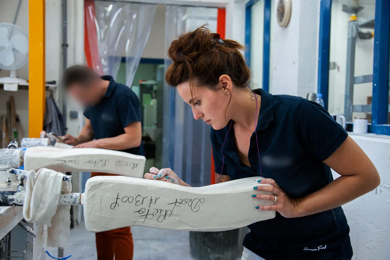 Dans un atelier, une ouvrière penchée sur un moulage en plâtre qu'elle retient de la main gauche. Derrière elle, un homme travaille sur une pièce semblable.