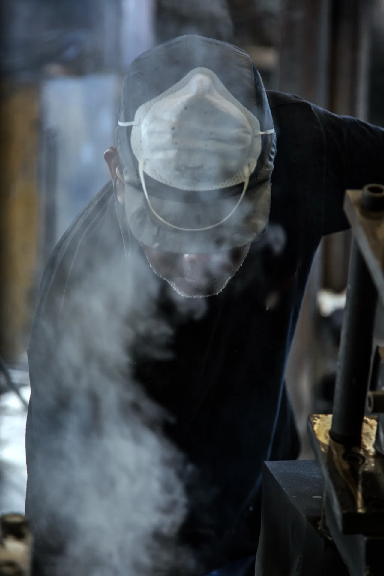 Un fondeur penché sur son poste, le visage pris dans un nuage de fumée, un masque en papier relevé sur la casquette.
