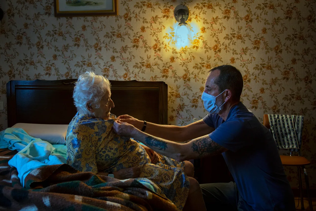 Dans une chambre au papier peint fleuri, un aide à domicile masqué, accroupi au bord du lit, ajuste le vêtement d'une femme très âgée assise.